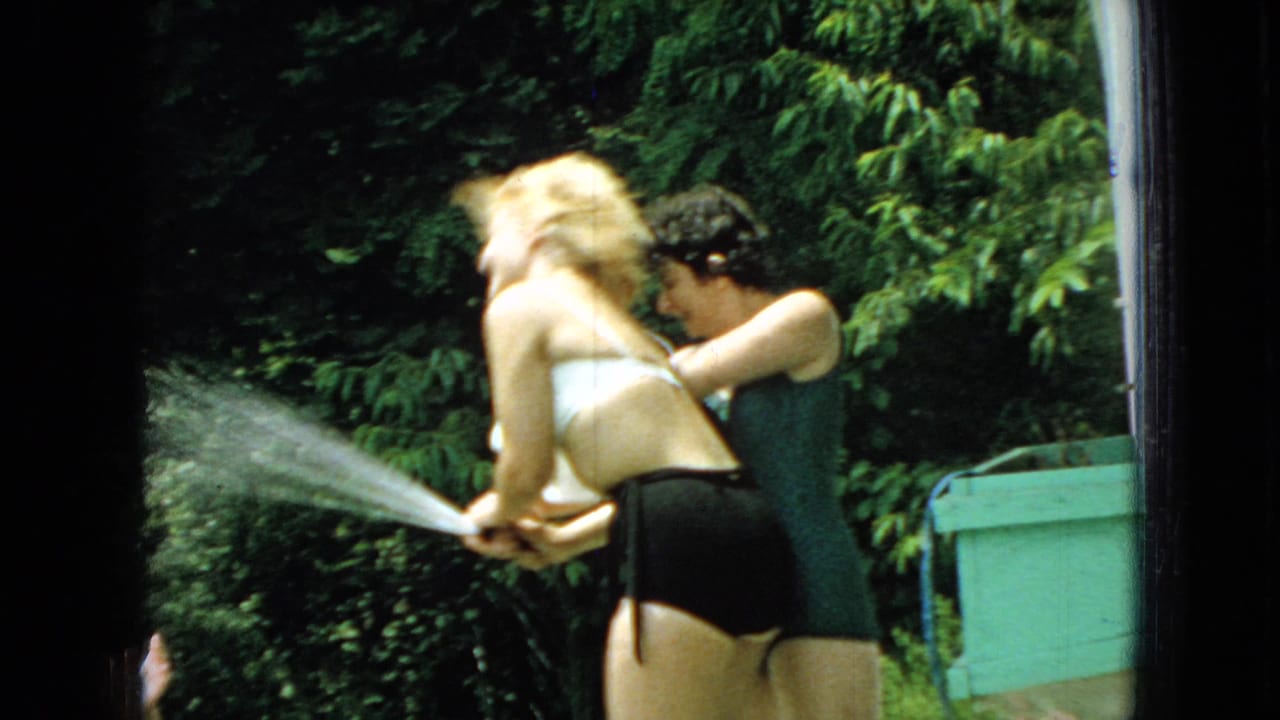 In metadata-supported 1965 Wellsburg, Iowa home-movie footage, two women in bathing suits playfully fight over a garden hose spraying across a green backyard.