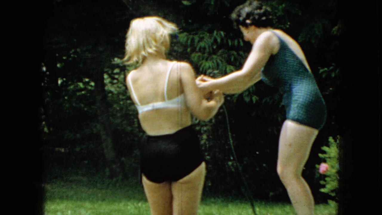 Two women in a sunlit backyard, one in a green one-piece swimsuit and one in a white top with dark bottoms, continue their playful tussle with a garden hose.
