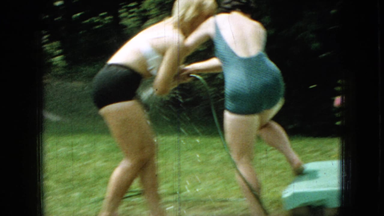 The handheld 8mm frame catches the women in motion as they bend and turn during the hose play, with grass and trees filling the backyard setting.