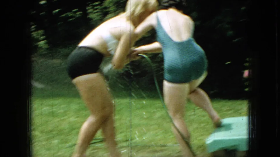 The handheld 8mm frame catches the women in motion as they bend and turn during the hose play, with grass and trees filling the backyard setting.