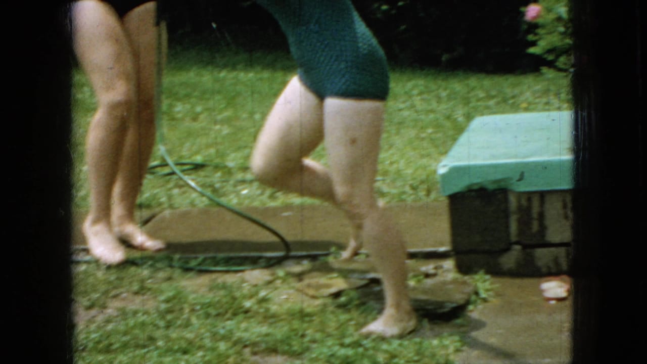In a 1965 Wellsburg, Iowa home-movie frame, barefoot family members in swimsuits stand near a hose and small yard structure on a green backyard lawn.