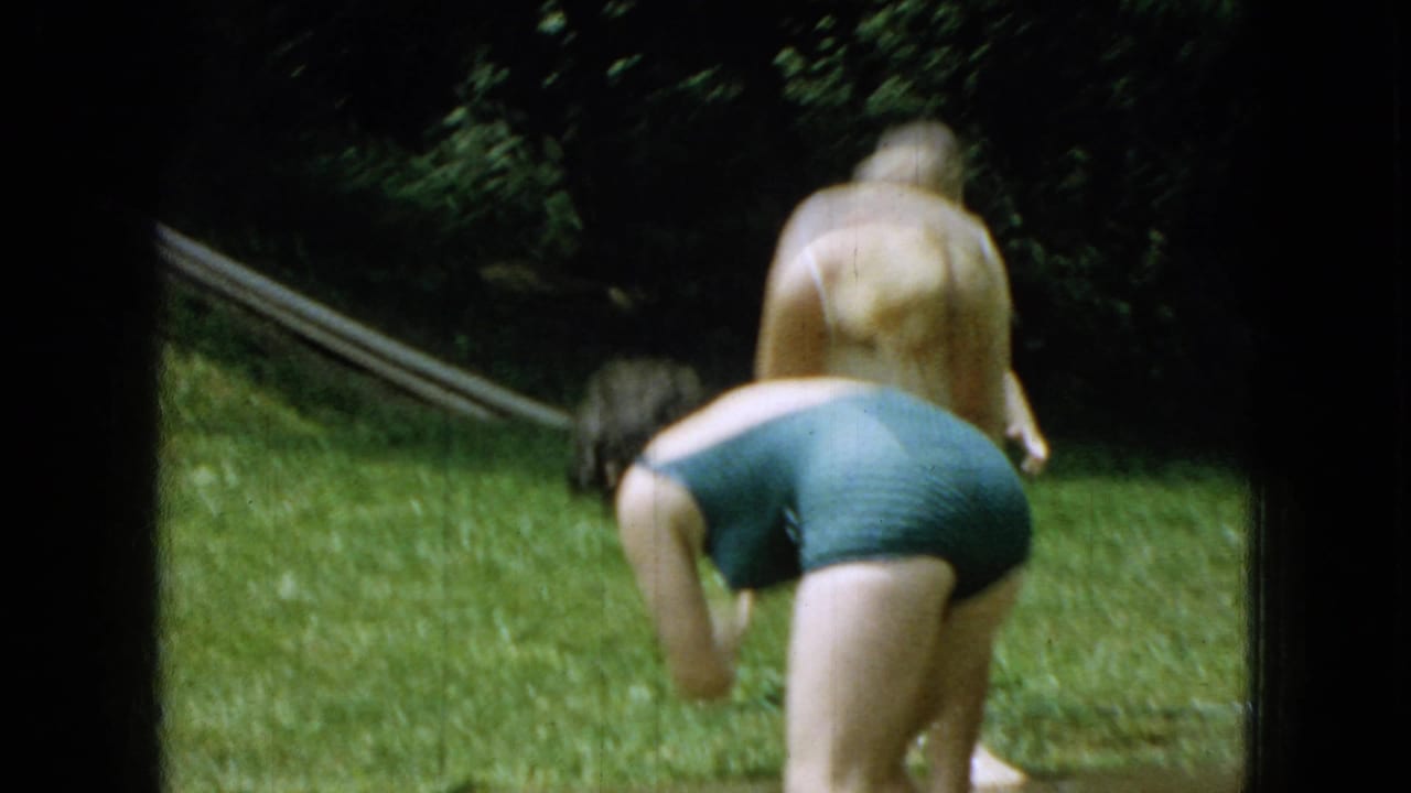 Two swimsuit-clad figures bend and move across a grassy backyard near a hose.
