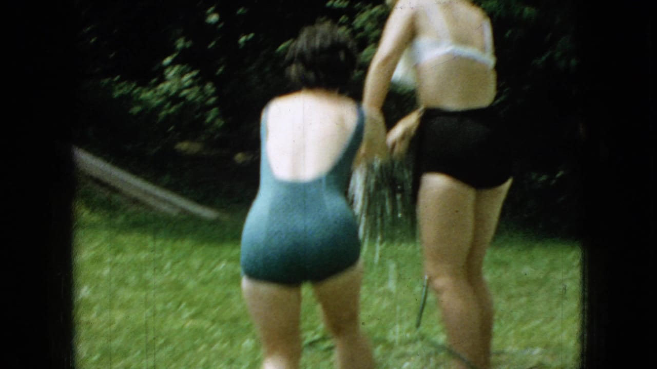 In this 1965 Wellsburg, Iowa home-movie frame, two swimsuit-clad family members stand on the grass while water sprays downward from a garden hose.