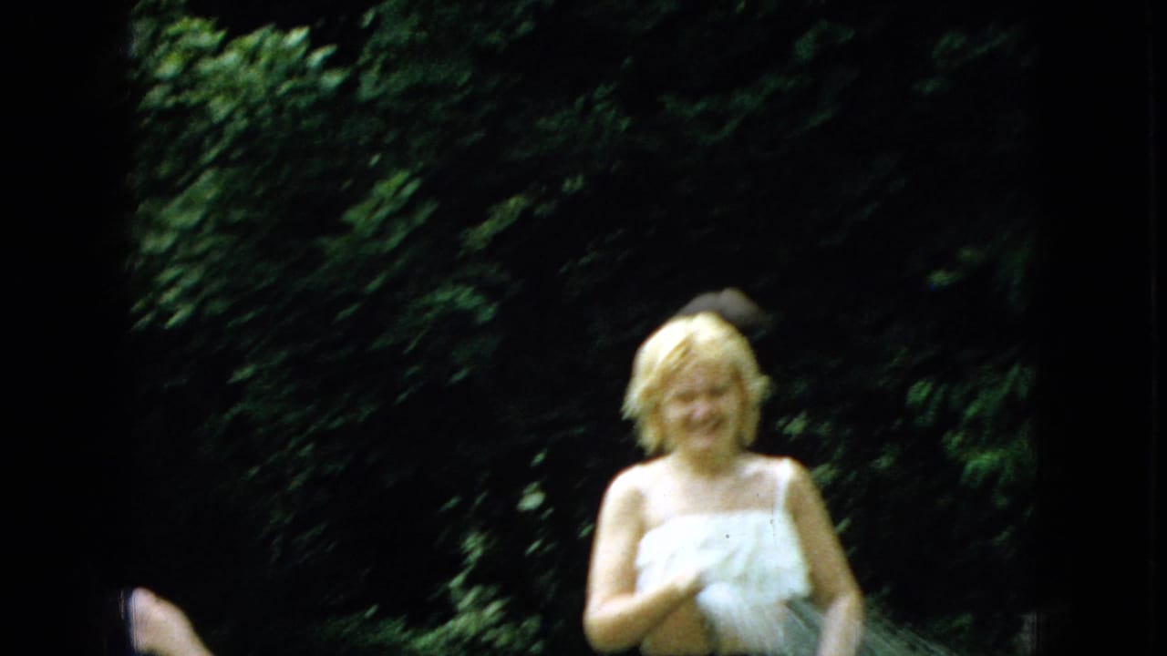 A 1965 Wellsburg, Iowa home-movie frame shows a blond family member holding a pale towel on the lawn, with dark greenery filling the background.