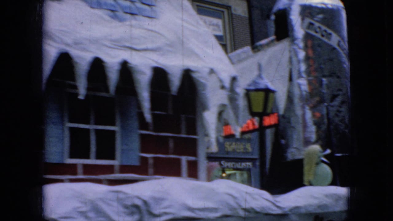 1965 Wellsburg, Iowa holiday parade frame showing a winter-decorated float with a brick-house facade, hanging icicles, and downtown storefront signage in soft 8mm focus.