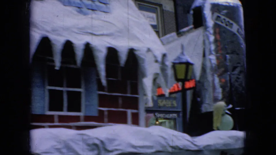 1965 Wellsburg, Iowa holiday parade frame showing a winter-decorated float with a brick-house facade, hanging icicles, and downtown storefront signage in soft 8mm focus.