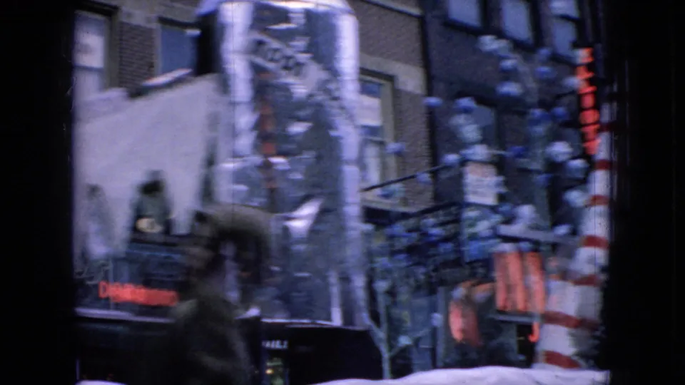 A holiday parade float with a tall candy cane and artificial snow passes in front of Wellsburg storefronts, with signage and building windows visible through handheld 8mm blur.