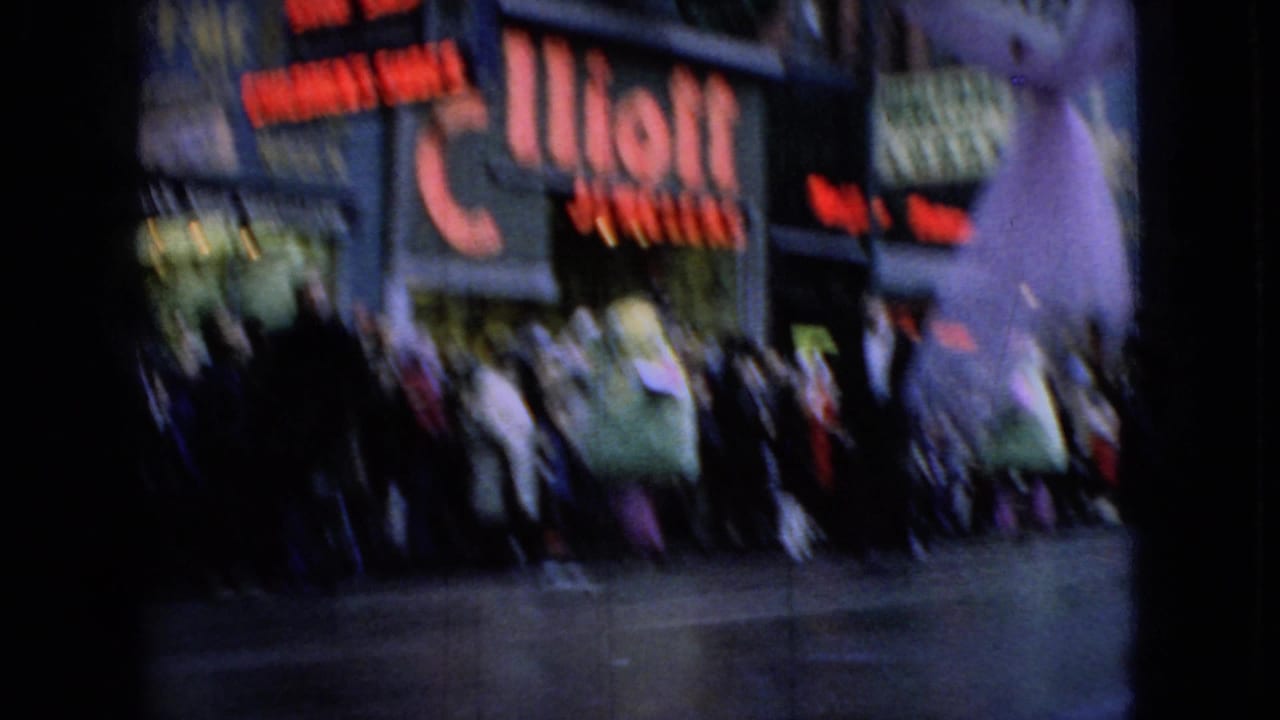 Handheld 8mm frame of a holiday parade crowd along a snowy commercial street, with an Elliott Jewelers sign and other storefront lettering visible behind the spectators.