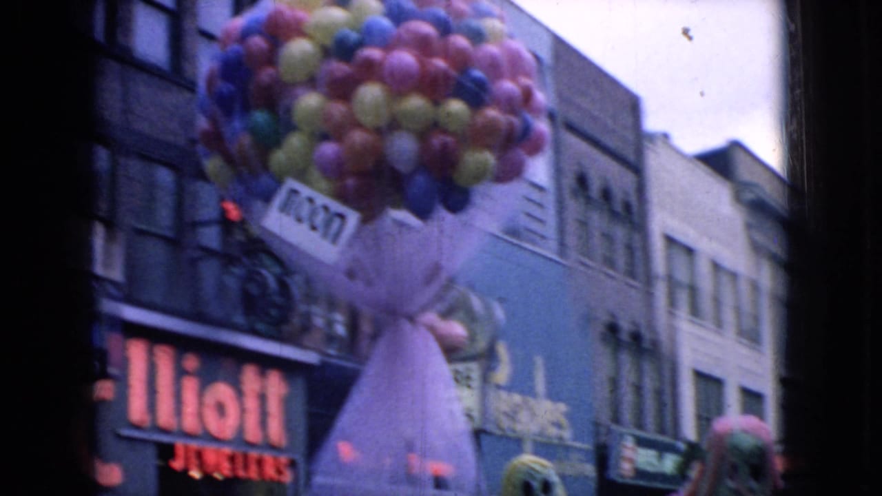 Colorful balloons rise above a holiday parade float on a Wellsburg commercial street, with the Elliott Jewelers sign and neighboring storefronts visible in the 1965 8mm frame.