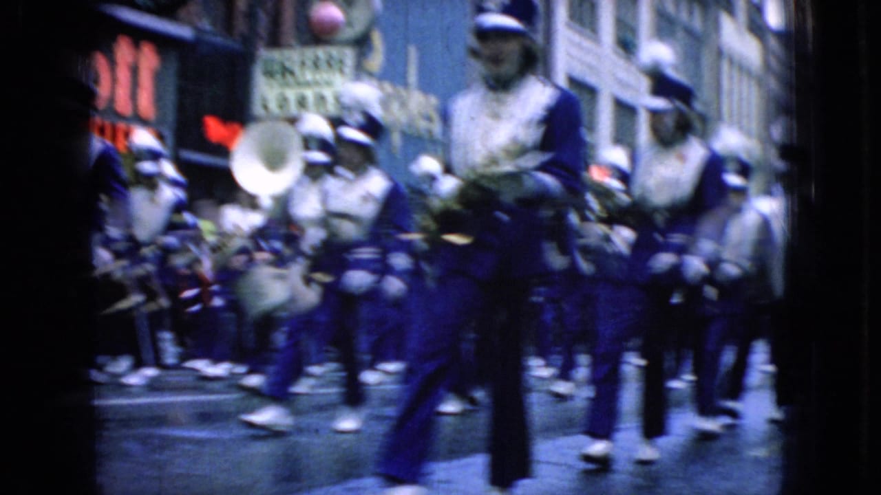 1965 Wellsburg, Iowa 8mm frame showing a marching band in blue-and-white uniforms passing storefronts while a crowd watches from the sidewalk.