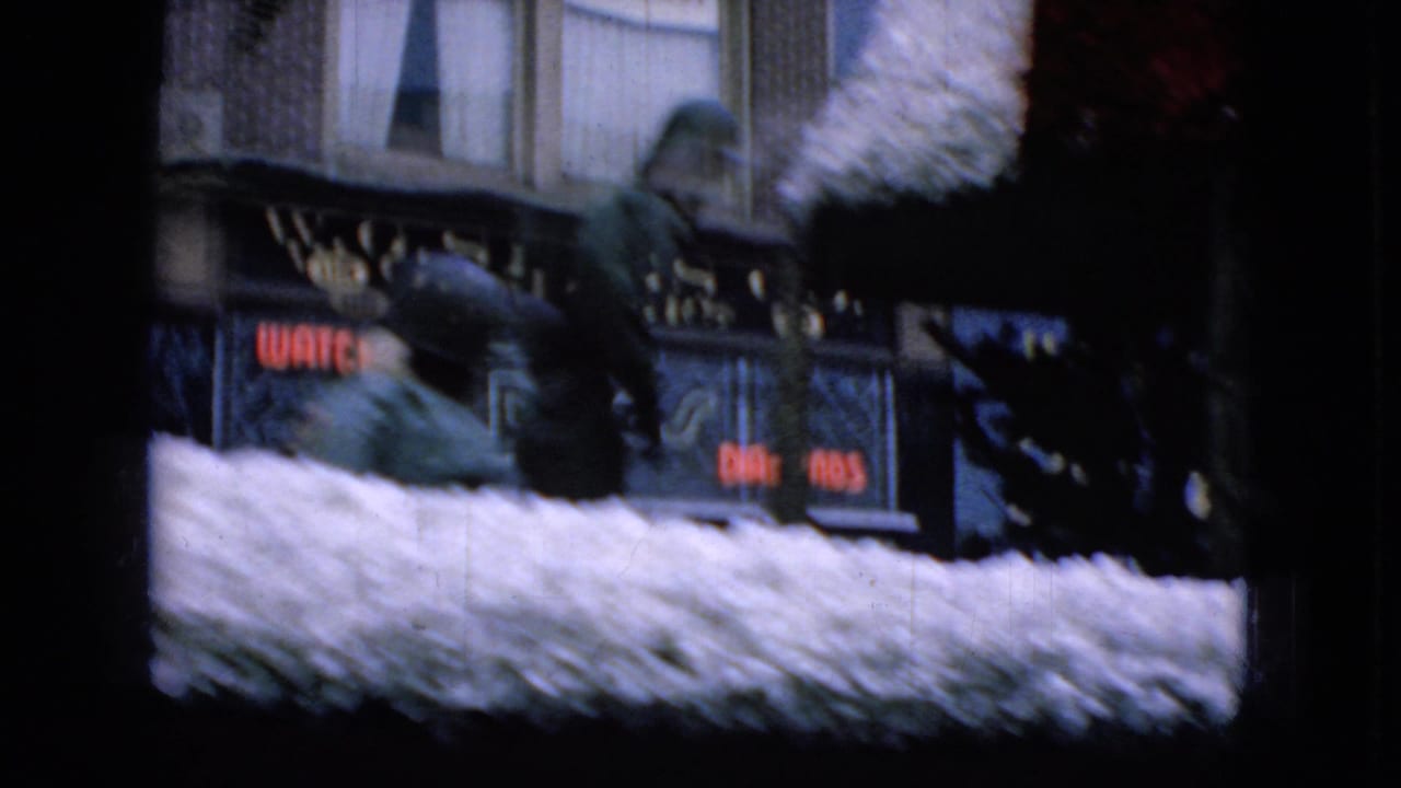 1965 Wellsburg, Iowa 8mm frame showing a white decorated parade float moving past commercial storefronts with partially readable signs in the background.