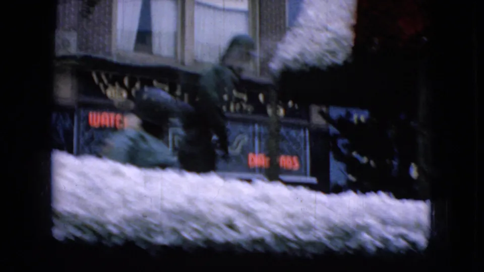 1965 Wellsburg, Iowa 8mm frame showing a white decorated parade float moving past commercial storefronts with partially readable signs in the background.