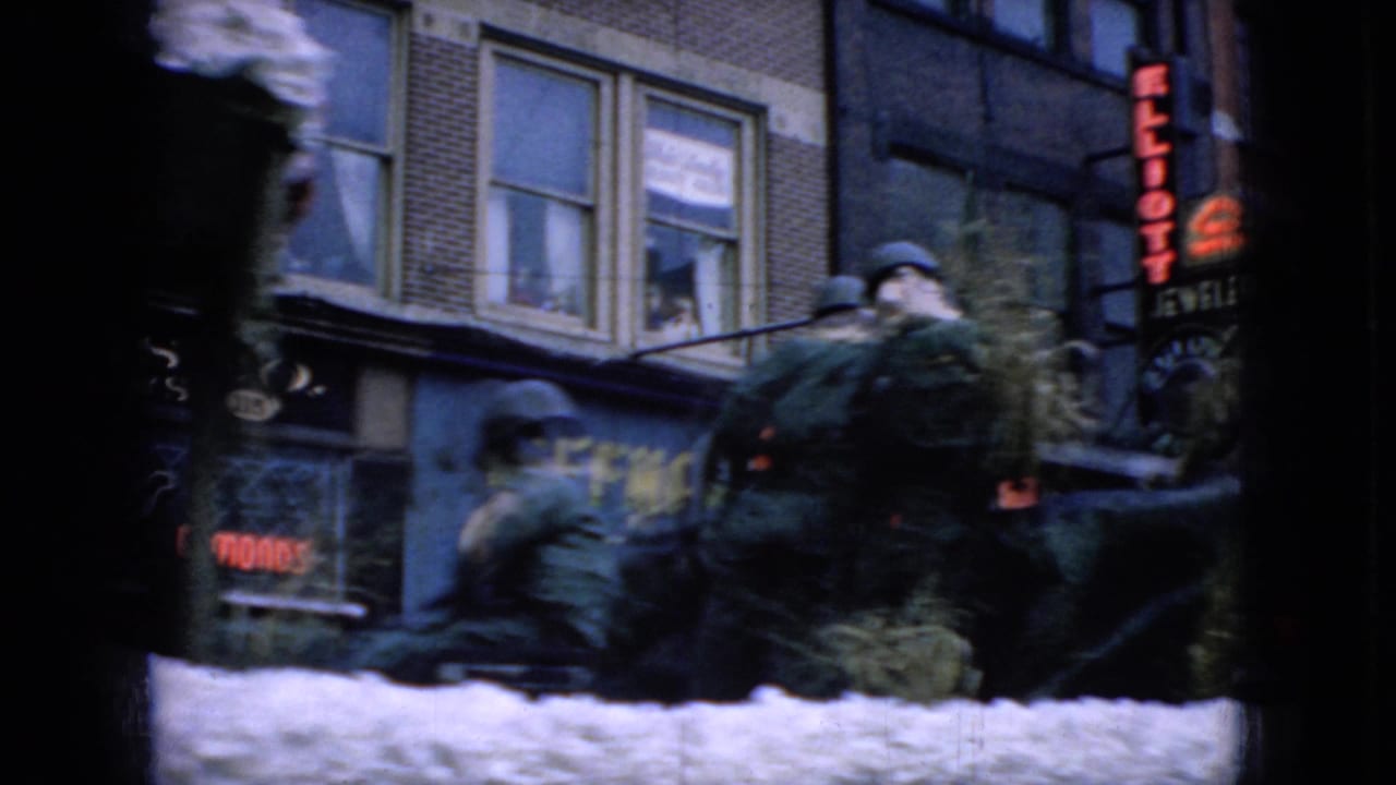 People ride or sit on a white parade float near storefront signs and a brick building.
