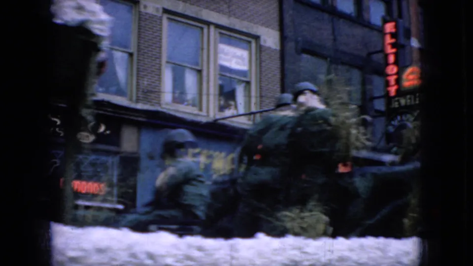1965 Wellsburg, Iowa 8mm frame showing riders or bundled figures on a white parade float beside downtown storefronts and visible commercial signage.