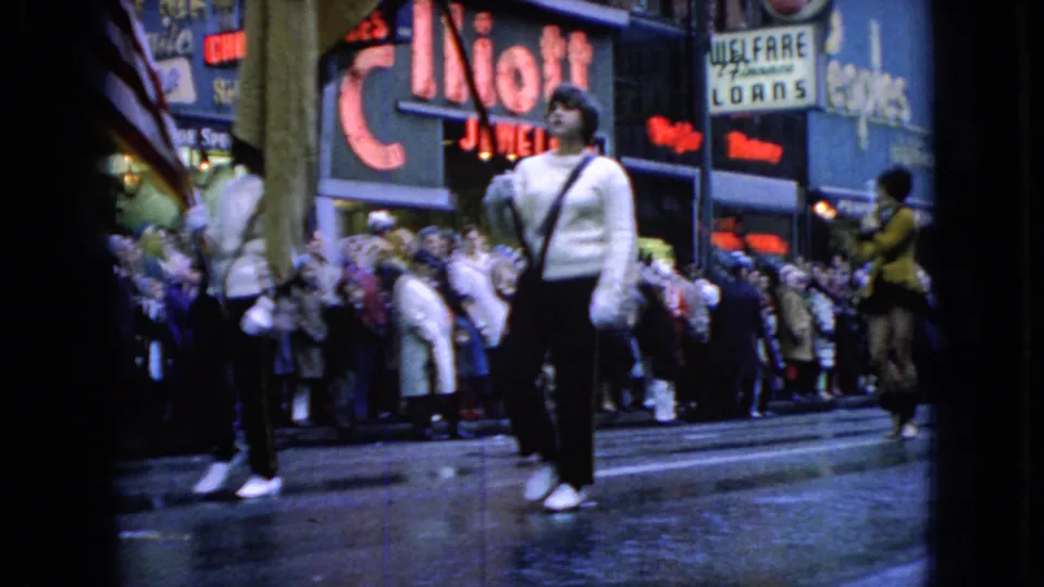 1965 Wellsburg, Iowa 8mm frame showing a white-uniformed parade marcher or drum major on a wet downtown street with spectators and commercial signs behind.