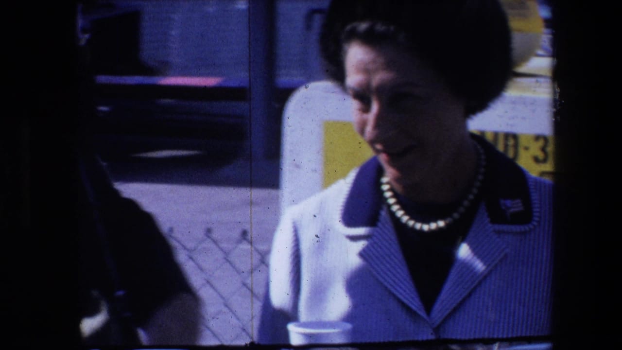 A woman in a white-and-blue striped jacket and pearl necklace appears outdoors in 1960s home-movie footage, with a dark foreground shape partly obscuring the left side.
