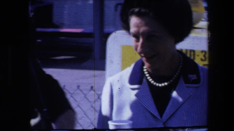 A woman in a white-and-blue striped jacket and pearl necklace appears outdoors in 1960s home-movie footage, with a dark foreground shape partly obscuring the left side.