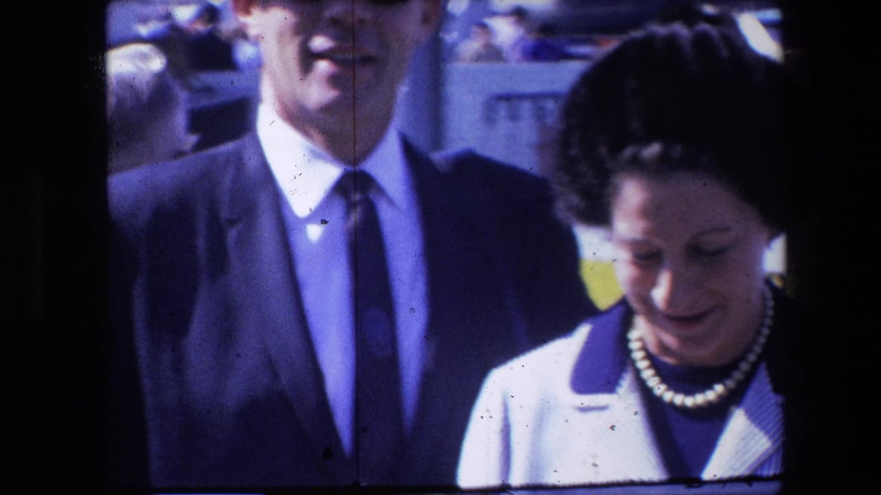 Man in a dark suit and woman in a striped jacket standing outdoors among nearby people.