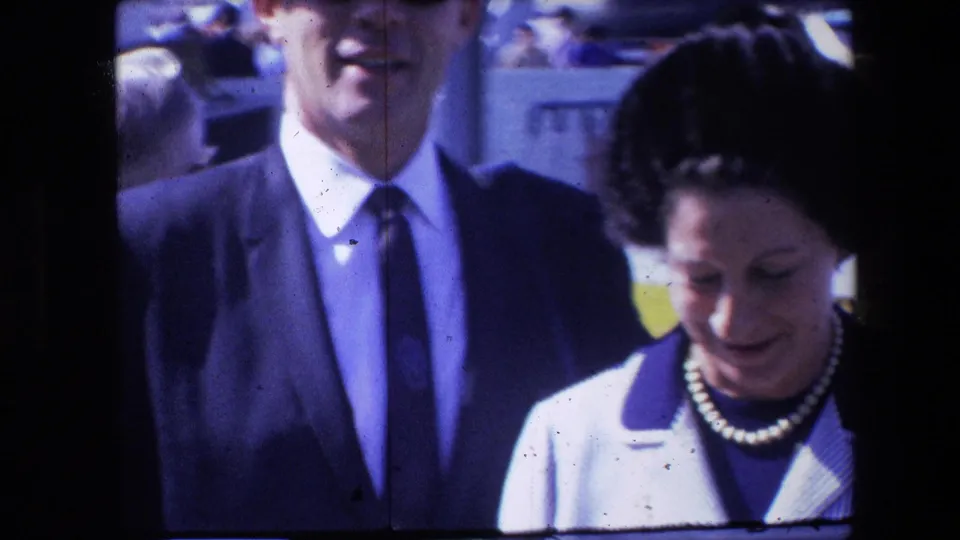 A man in a dark suit and tie stands beside a woman in a striped jacket and pearl necklace, with nearby people and a partial yellow background element visible.