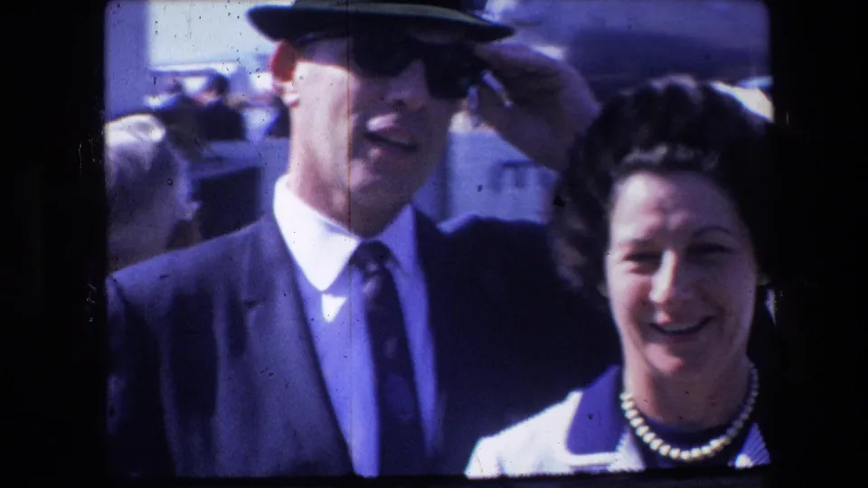 A man in a dark suit and tie raises or adjusts a brimmed hat while a woman in a striped jacket smiles beside him in bright outdoor home-movie footage.
