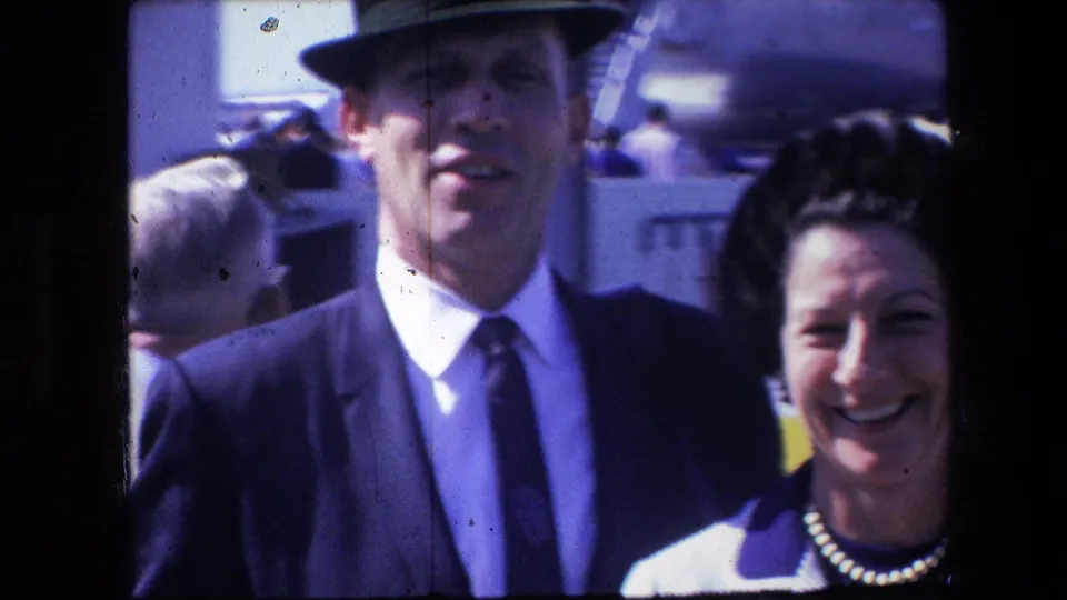 A smiling adult couple appears outdoors in 1960s home-movie footage, the man wearing a suit and hat and the woman wearing a striped jacket with a pearl necklace.