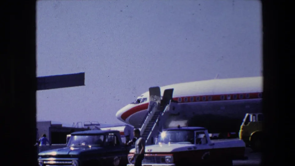 A 1960s 8mm frame shows a World Airways passenger aircraft on a bright airport apron, with mobile stairs, pickup trucks, service vehicles, and several people near the plane.