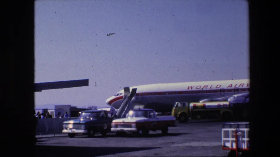 The final frame keeps the World Airways aircraft and stairs in view while pickup trucks and a service vehicle sit on the apron, with small groups of people near the ramp area.