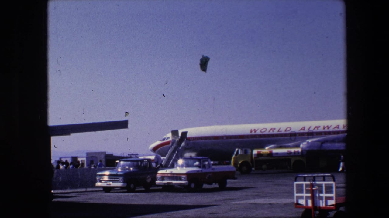 1967 Amsterdam airport home-movie frame showing a World Airways aircraft parked on the tarmac with boarding stairs, a pickup, a service truck and people near the plane.