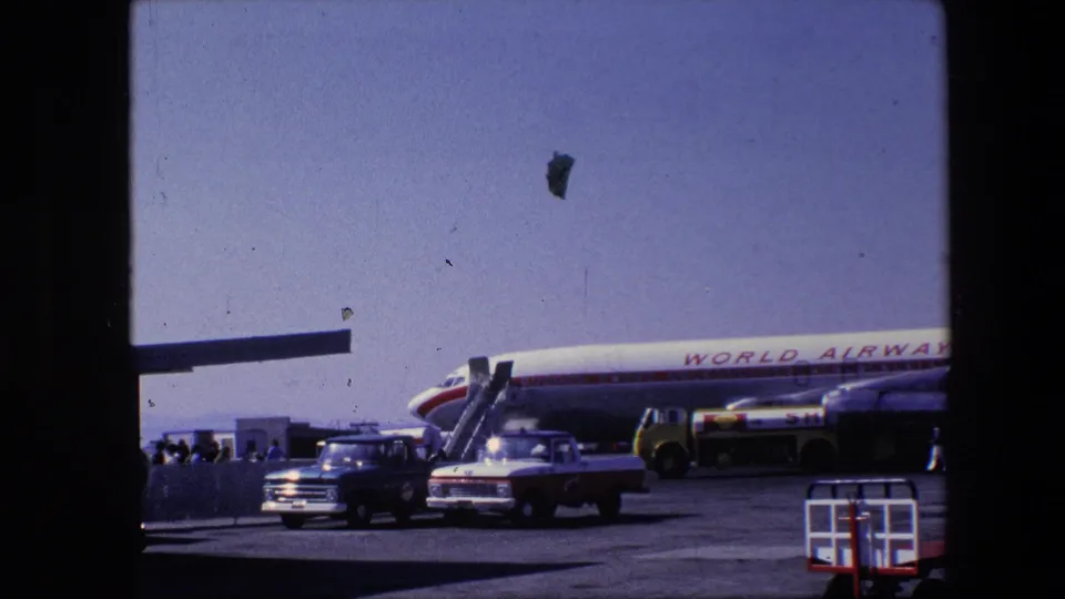1967 Amsterdam airport home-movie frame showing a World Airways aircraft parked on the tarmac with boarding stairs, a pickup, a service truck and people near the plane.