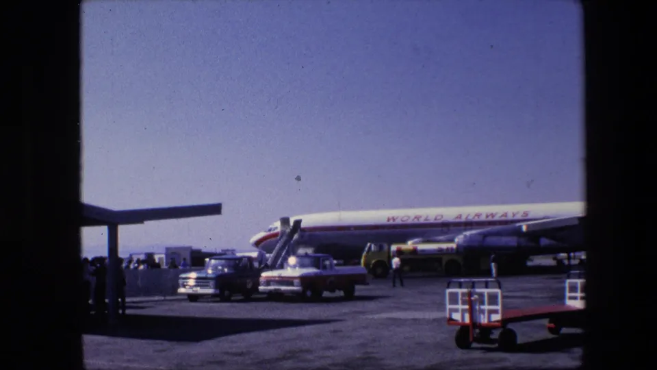 1967 Amsterdam airport home-movie view across the tarmac toward a World Airways aircraft, with a pickup, service truck, boarding stairs and small figures visible.