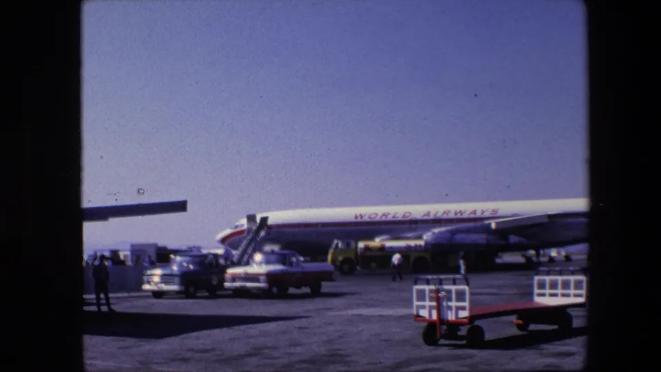 1967 Amsterdam airport frame showing a World Airways aircraft on the tarmac with service vehicles, baggage carts and a few people walking or standing nearby.
