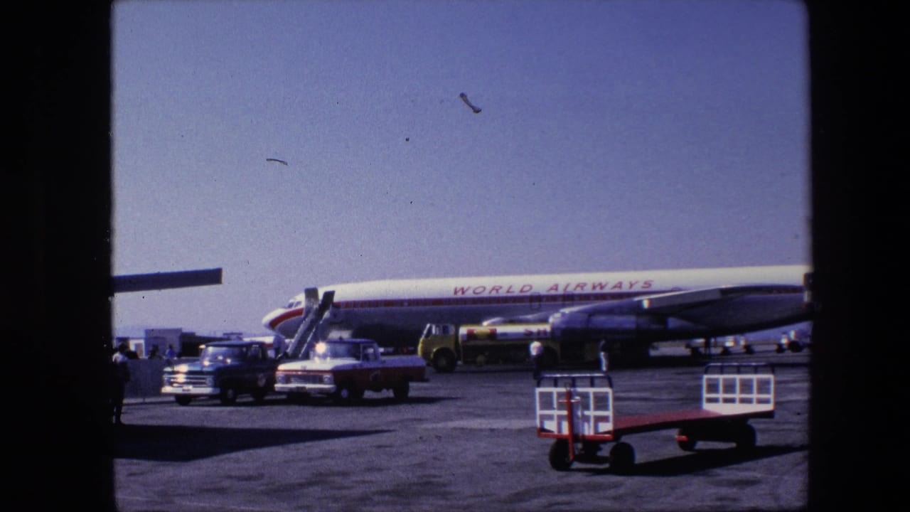 1960s 8mm frame showing a World Airways passenger jet on an airport tarmac with stair equipment, vintage vehicles, and ground-service activity under a clear sky.