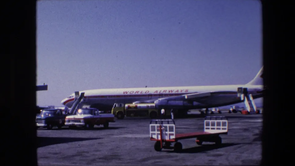 A stationary 8mm view of a parked World Airways commercial airliner, with ground-service vehicles, boarding stairs, and small figures on the airport apron.