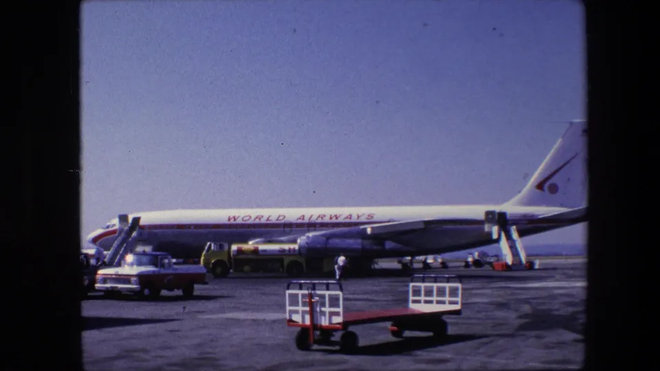 The aircraft remains parked in profile, with the World Airways name along the fuselage, tail markings, service stairs, vintage vehicles, and small tarmac carts in view.