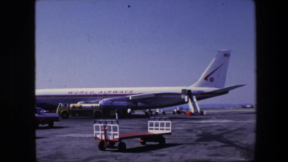 Wide 8mm frame of a World Airways jet parked across the airport apron, with ground carts in the foreground, stair equipment near the fuselage, and people working nearby.