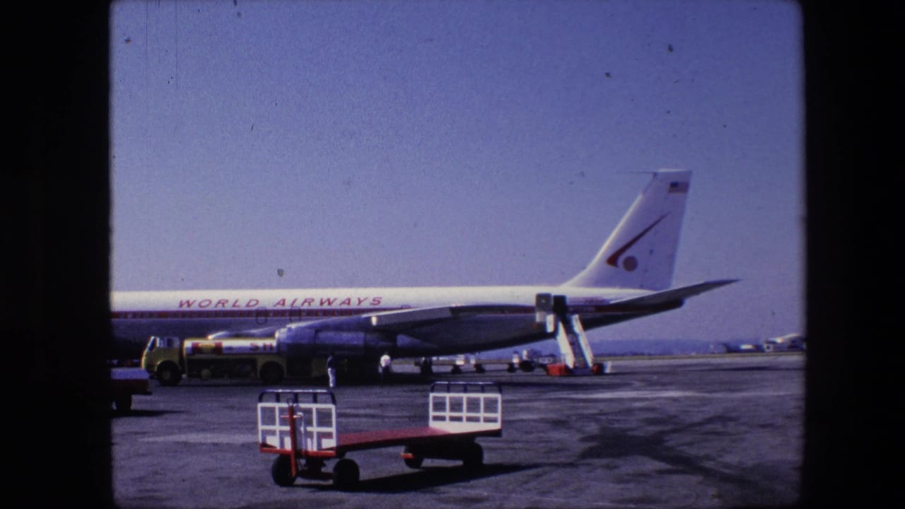 1967 archival home-movie frame of a World Airways commercial aircraft on an airport tarmac, with a mobile stairway, service vehicle, and baggage cart visible.