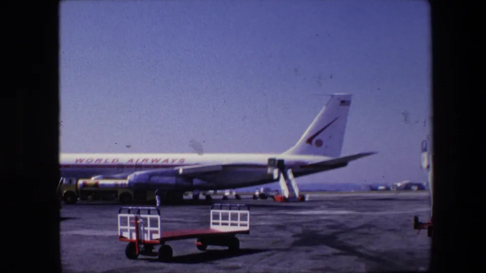 Side view of a World Airways jet on the tarmac in 1967 archival 8mm footage, with ground service equipment and runway pavement in view.