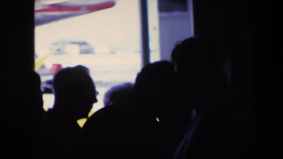 A group of mostly silhouetted travelers appears in the terminal foreground while a commercial airplane sits or moves outside the Amsterdam airport window.