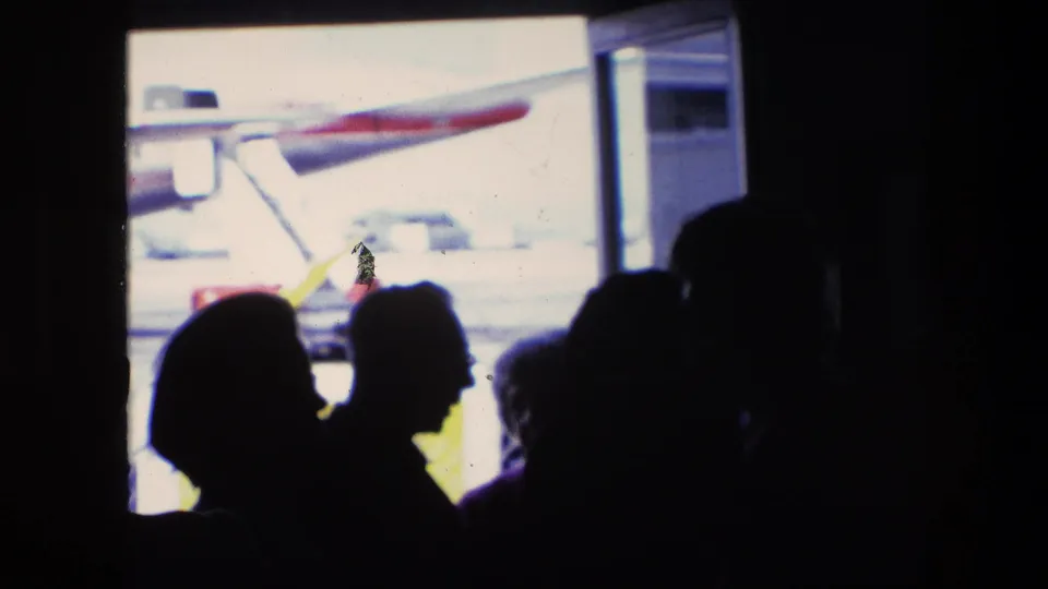 Travelers in profile form dark silhouettes inside the terminal, with a red-and-white airplane visible through the window in this 1967 Amsterdam airport footage.