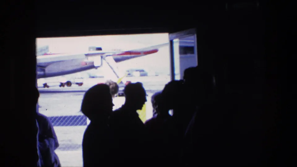 1967 home-movie airport scene with passengers in silhouette near a bright doorway and an aircraft visible on the tarmac.