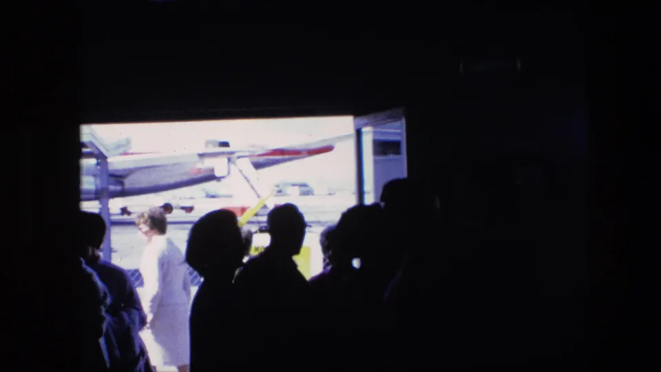 Silhouetted travelers stand inside a dim airport space as a person crosses the bright tarmac near the visible aircraft.