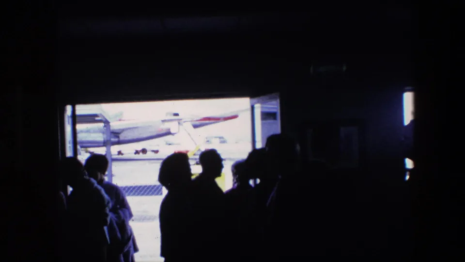 Home-movie view from a shadowed airport or aircraft interior toward passengers and a visible airplane on the sunlit tarmac.
