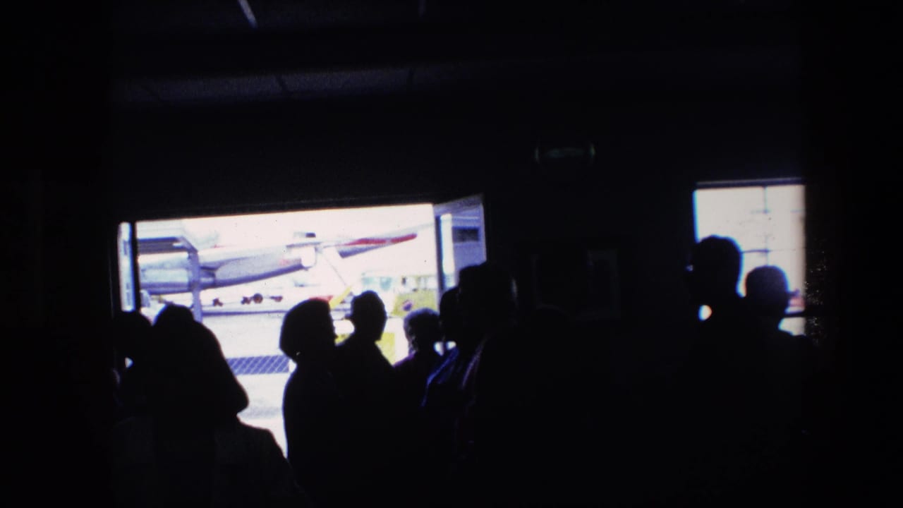 Backlit travelers stand in a dark airport terminal area, with a bright doorway revealing part of an aircraft on the tarmac in footage identified as 1967 airport travel.