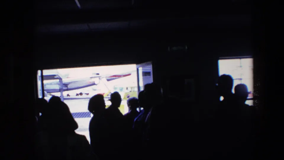 Backlit travelers stand in a dark airport terminal area, with a bright doorway revealing part of an aircraft on the tarmac in footage identified as 1967 airport travel.