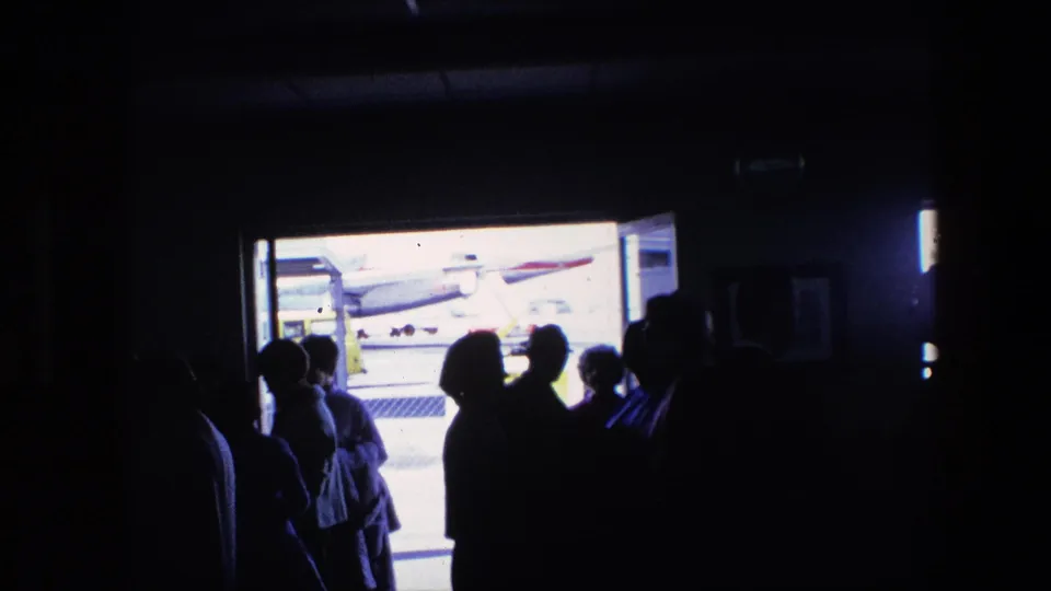Passengers appear in silhouette inside a dark terminal space while a commercial aircraft is visible outside through the bright opening, consistent with supplied 1967 airport footage.