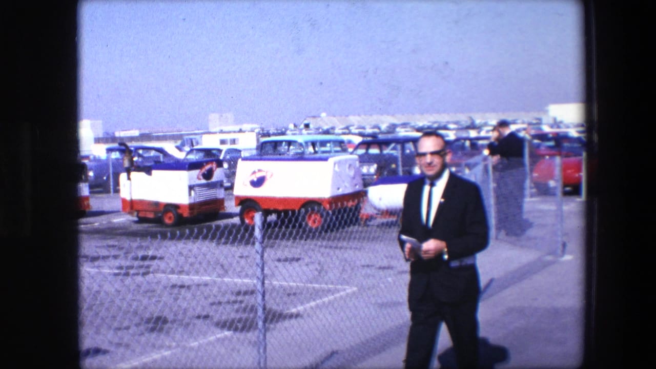 In the supplied 1967 clip, a man in a dark suit and glasses stands in a parking lot holding papers while red-and-white NASA-marked vehicles and classic cars sit behind a chain-link fence.