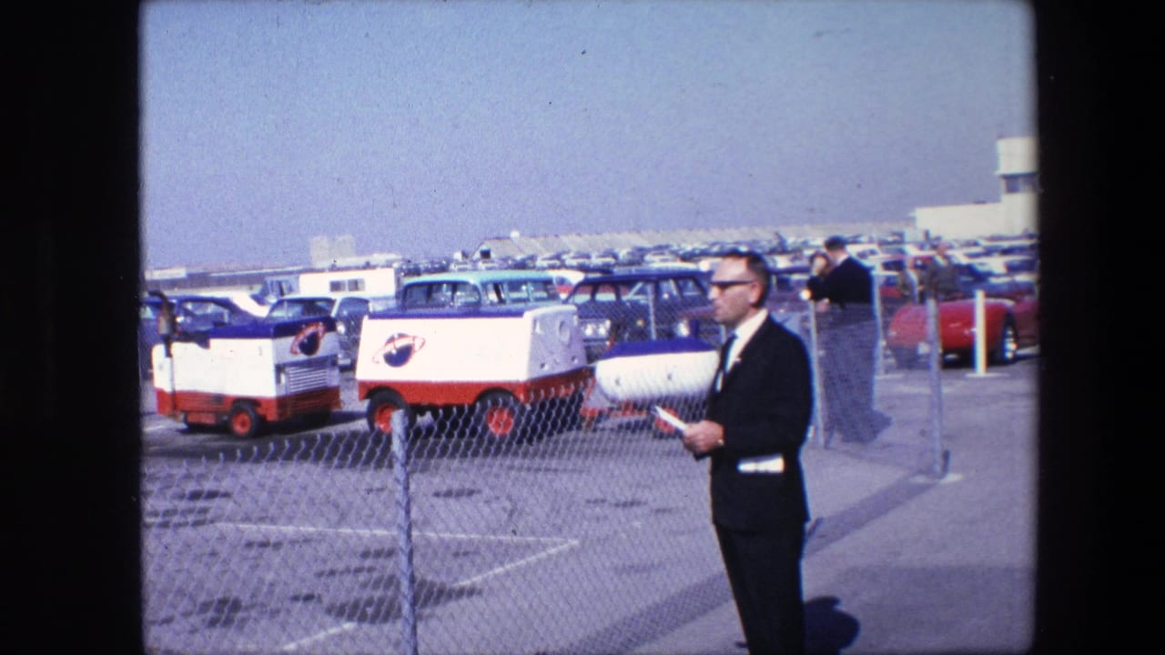 Man in dark suit seen in profile beside a fenced parking area with NASA-marked vehicles.