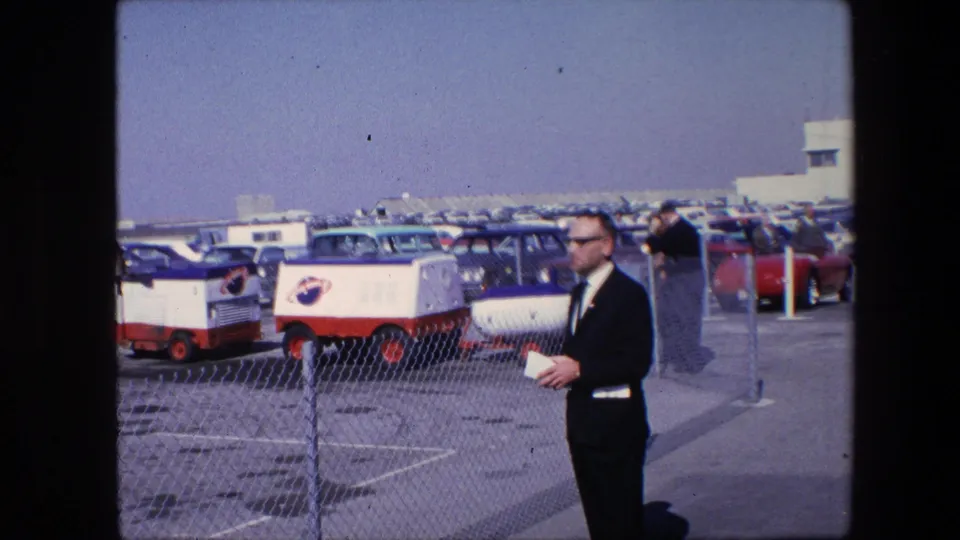 A man in a dark suit and sunglasses or dark glasses holds papers near a chain-link fence as red-and-white NASA-marked vehicles and rows of parked cars fill the background.