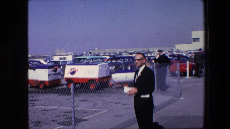 The suited man faces slightly toward the camera while holding papers, with a chain-link fence, NASA-marked red-and-white vehicles, classic cars, and a clear sky behind him.