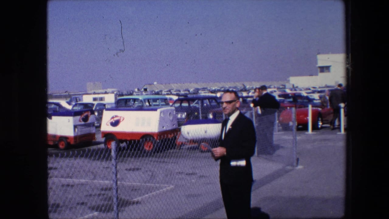 1967 8mm frame showing a suited man in sunglasses near a chain-link fence, with older cars and red-and-white service carts in a sunny United States parking lot.